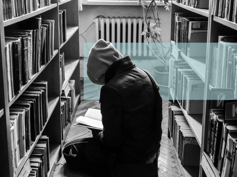 Sort-hvid foto af en person med hætte, der læser på gulvet i et bibliotek.